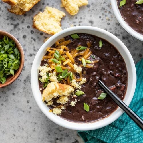 Two bowls of Smoky Black Bean Soup with various toppings, next to a bowl of sliced green onion and a crumbled corn muffin.