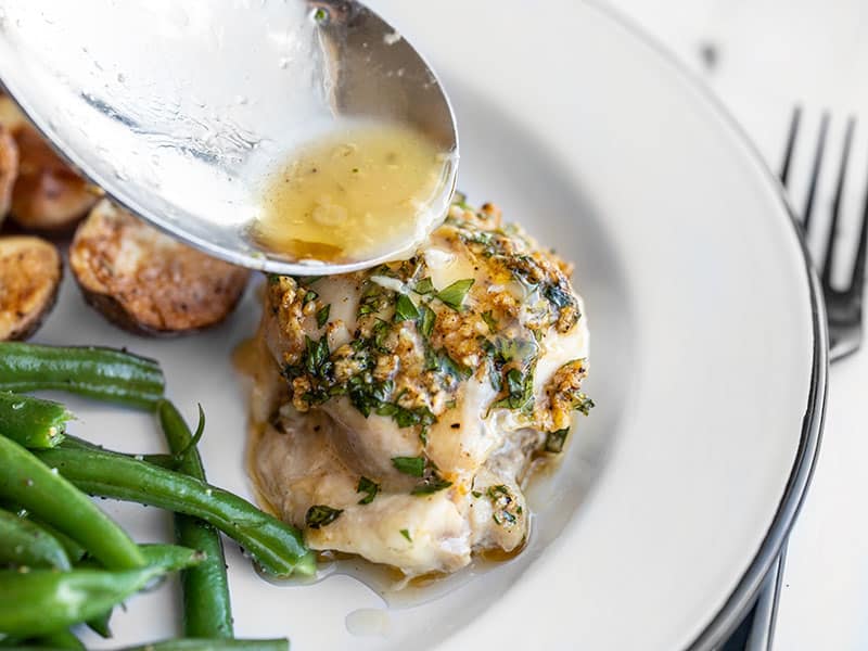 Juices being drizzled over garlic butter baked chicken thighs on a plate with potatoes and green beans