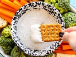 A cracker being dipped into the whipped cottage cheese dip, the platter of vegetables and crackers in the background