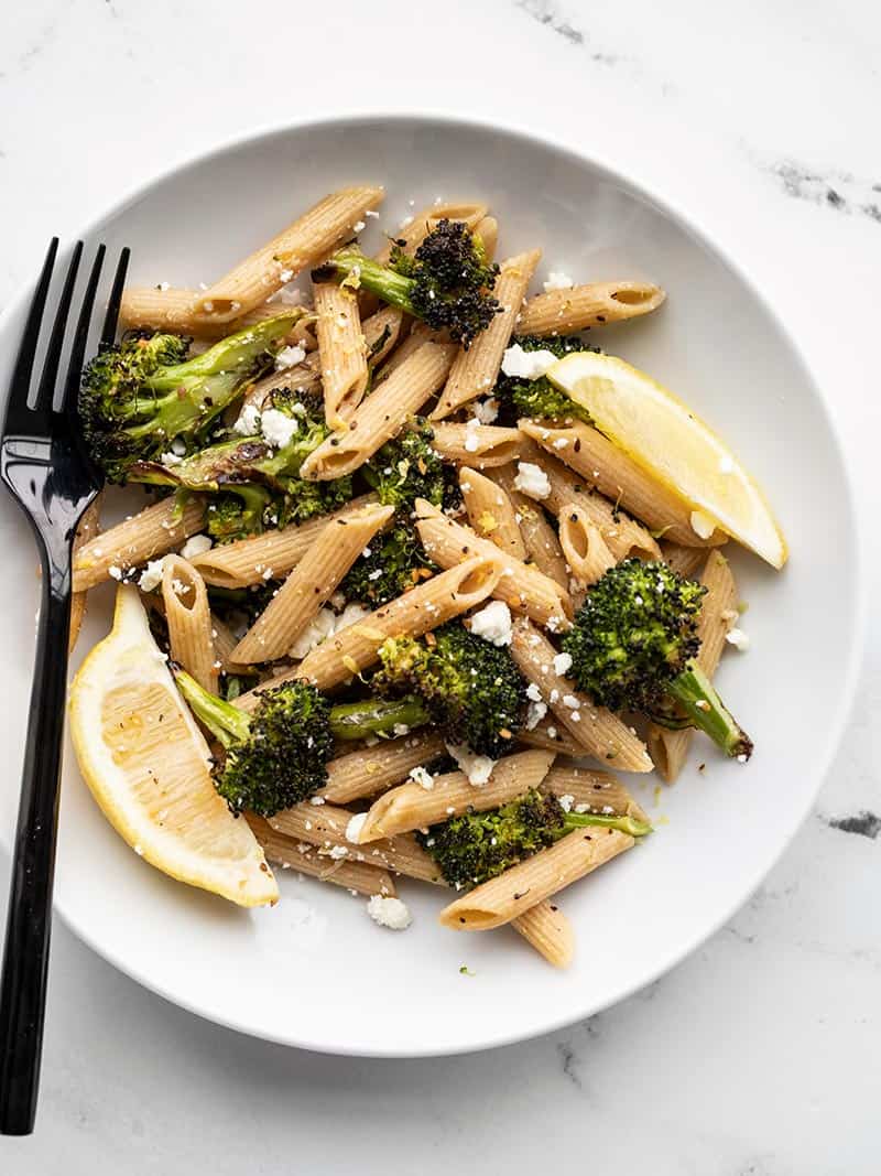 A plate with roasted broccoli pasta, lemon wedges, feta, and a black fork on the left