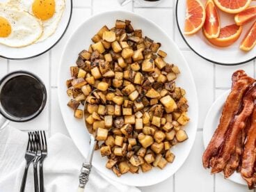 A platter full of roasted breakfast potatoes with eggs, coffee, oranges, and bacon on the side