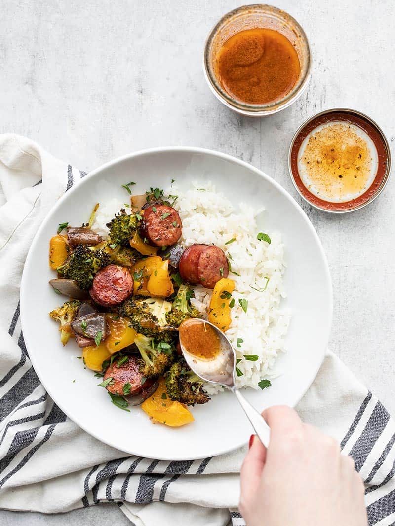 A bowl of rice with smoky roasted sausage and vegetables, vinaigrette being drizzled on top with a spoon.