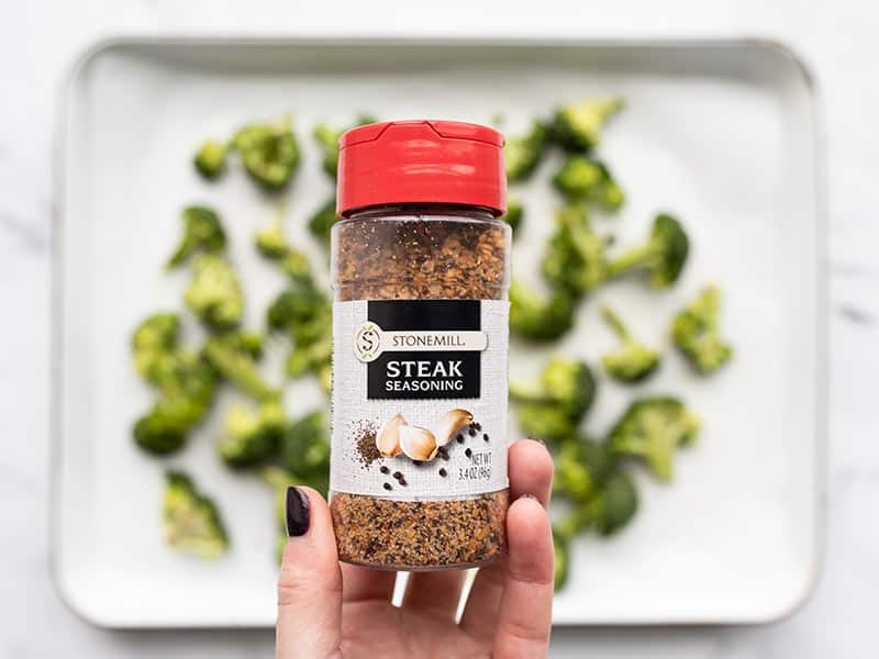 Steak seasoning bottle held over sheet pan with broccoli