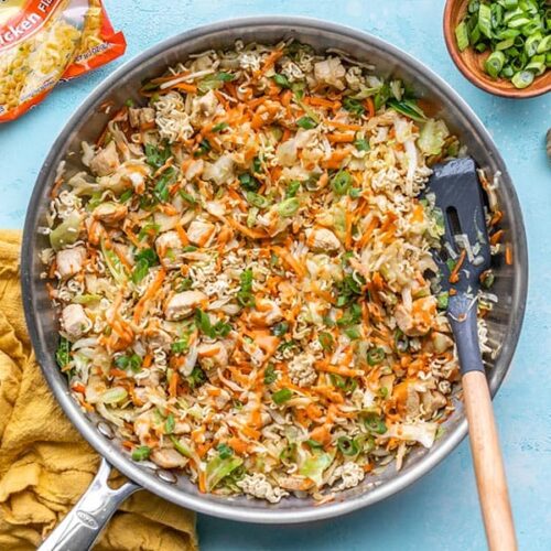 A skillet full of Crunchy Chicken Ramen Stir fry with a spatula in the side and a package of ramen on the left