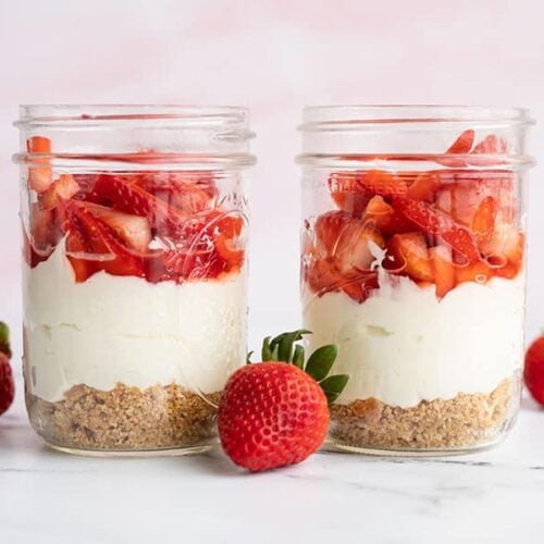 Two jars of no bake strawberry cheesecake side by side with whole strawberries on the side