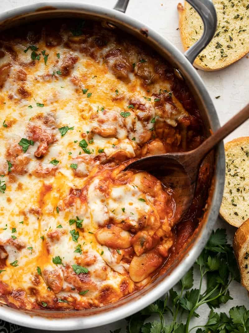 Close up overhead view of a skillet full of white beans with mushrooms and marinara and a wooden spoon scooping some out.