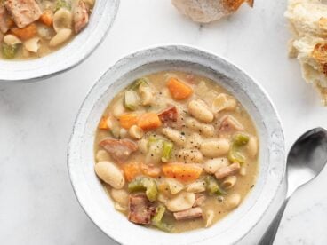 Two bowls of chunky ham and bean soup with chunks of bread on the side