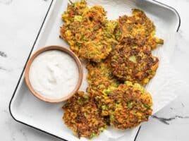 Overhead view of a white tray lined with paper towel, with vegetable fritters and a small bowl of garlic herb dipping sauce