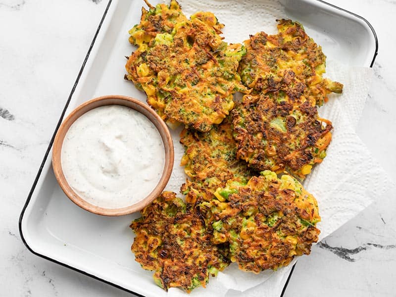 Overhead view of a white tray lined with paper towel, with vegetable fritters and a small bowl of garlic herb dipping sauce