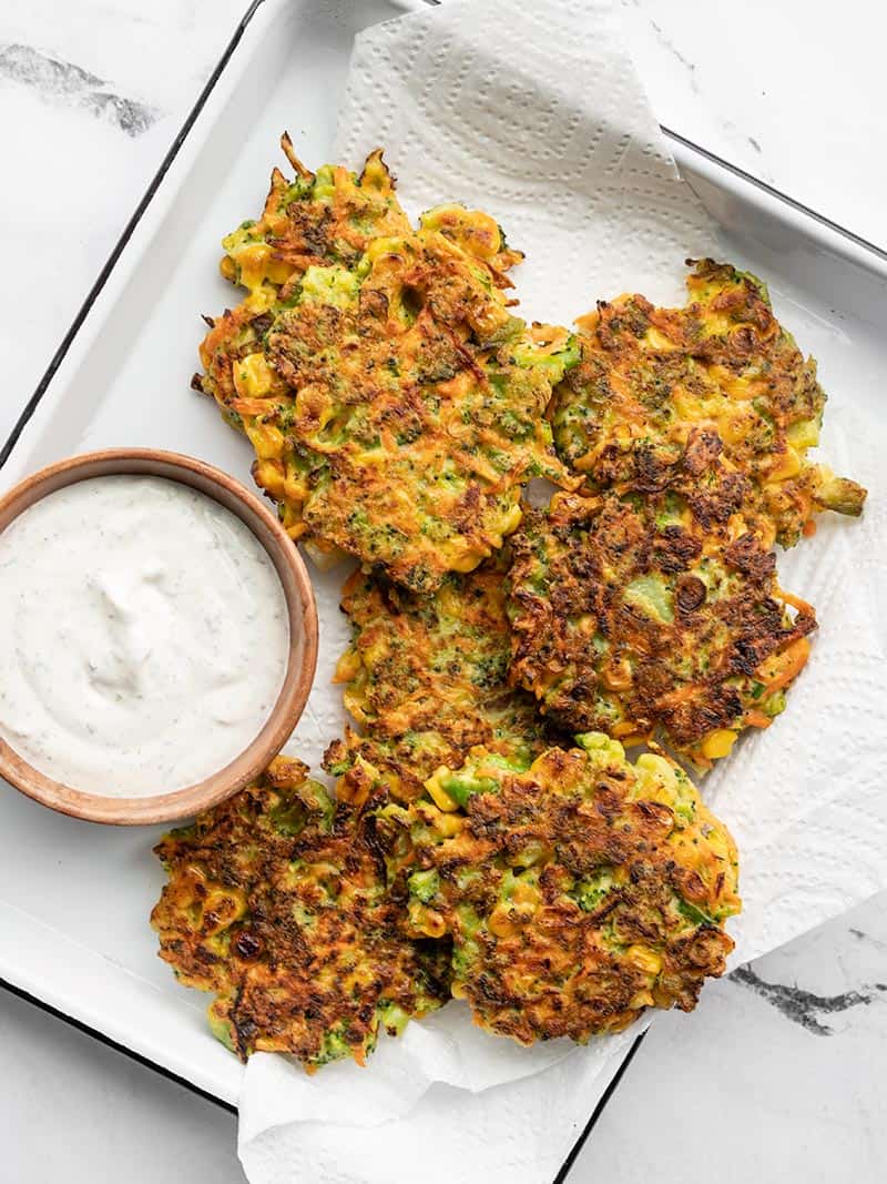 Overhead of vegetable fritters on a paper towel lined white tray, with a bowl of garlic herb dipping sauce on the side