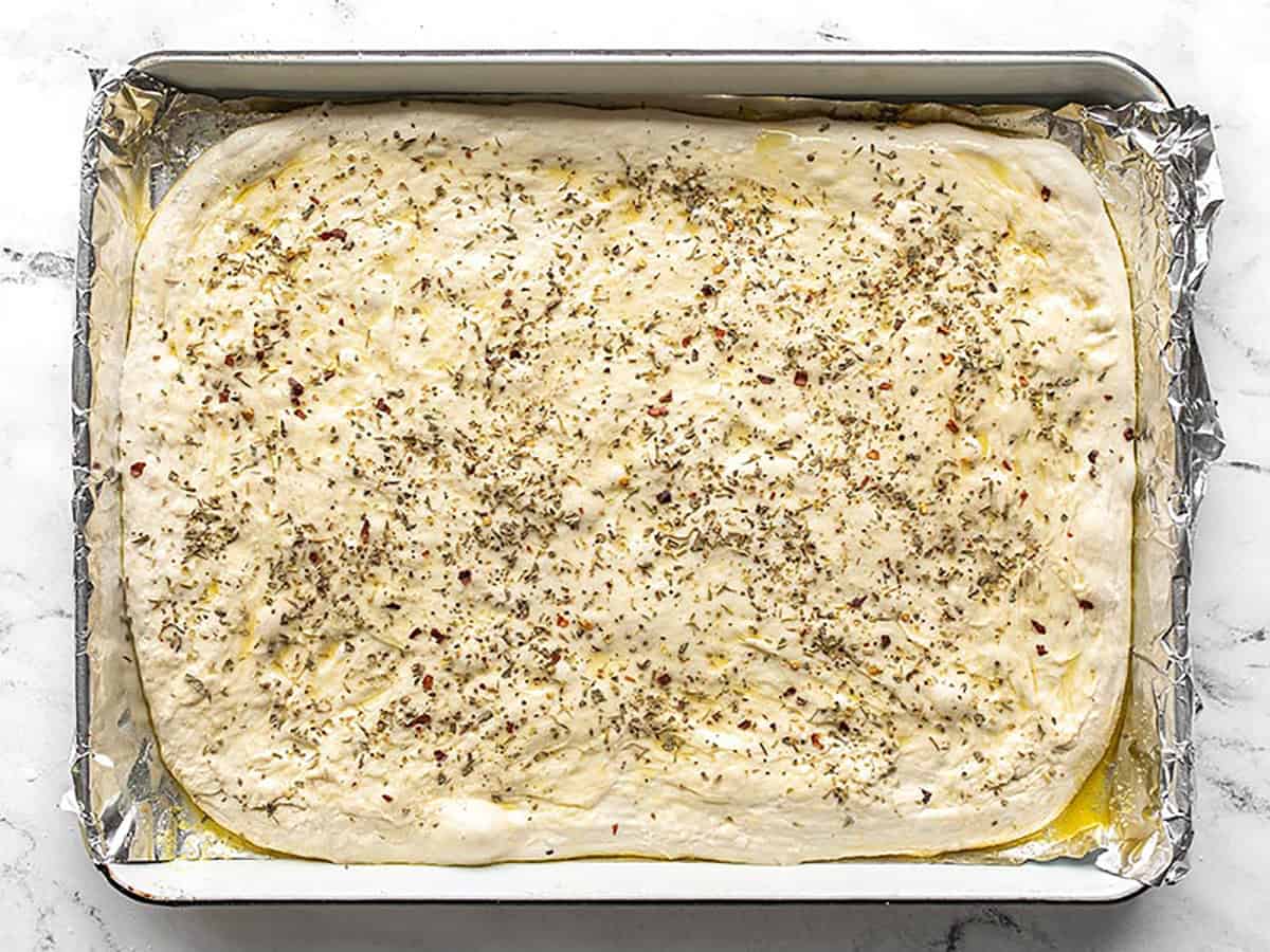 Focaccia dough stretched out onto the baking sheet and coated with herbs. 