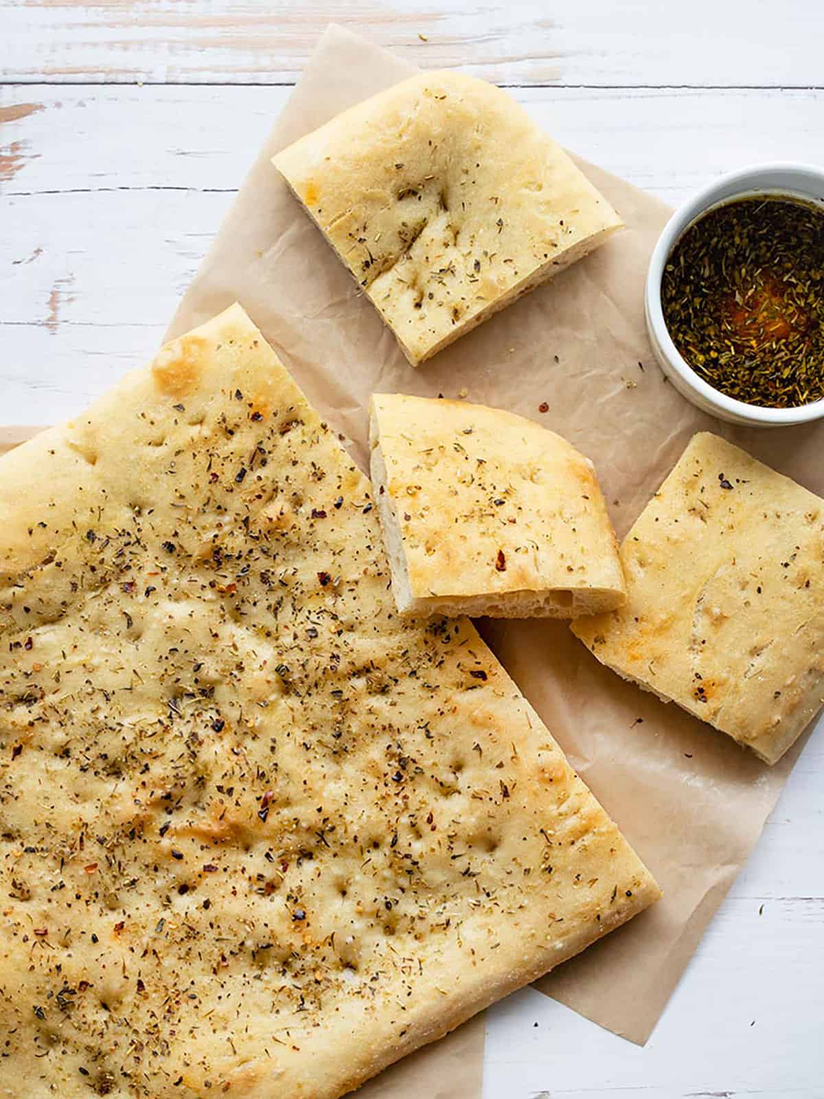 Overhead view of a loaf of focaccia bread on parchment with a few pieces cut and a bowl of dipping oil.