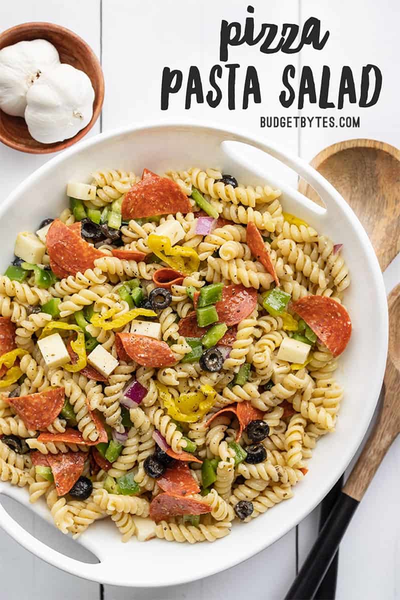 Overhead view of a serving bowl full of pizza pasta salad with wooden utensils on the side. Title text at the top.