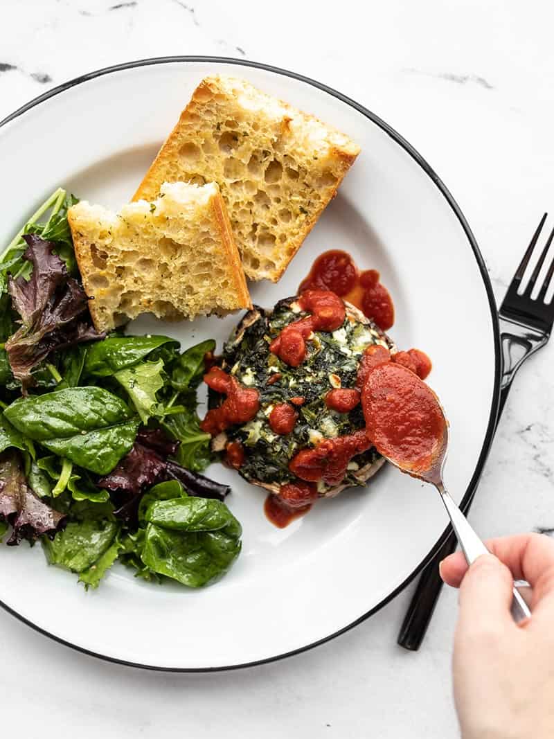 Marinara being spooned over a spinach stuffed portobello on a plate with salad and garlic bread