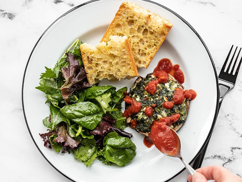 Marinara being spooned over a spinach stuffed portobello on a plate with salad and garlic bread