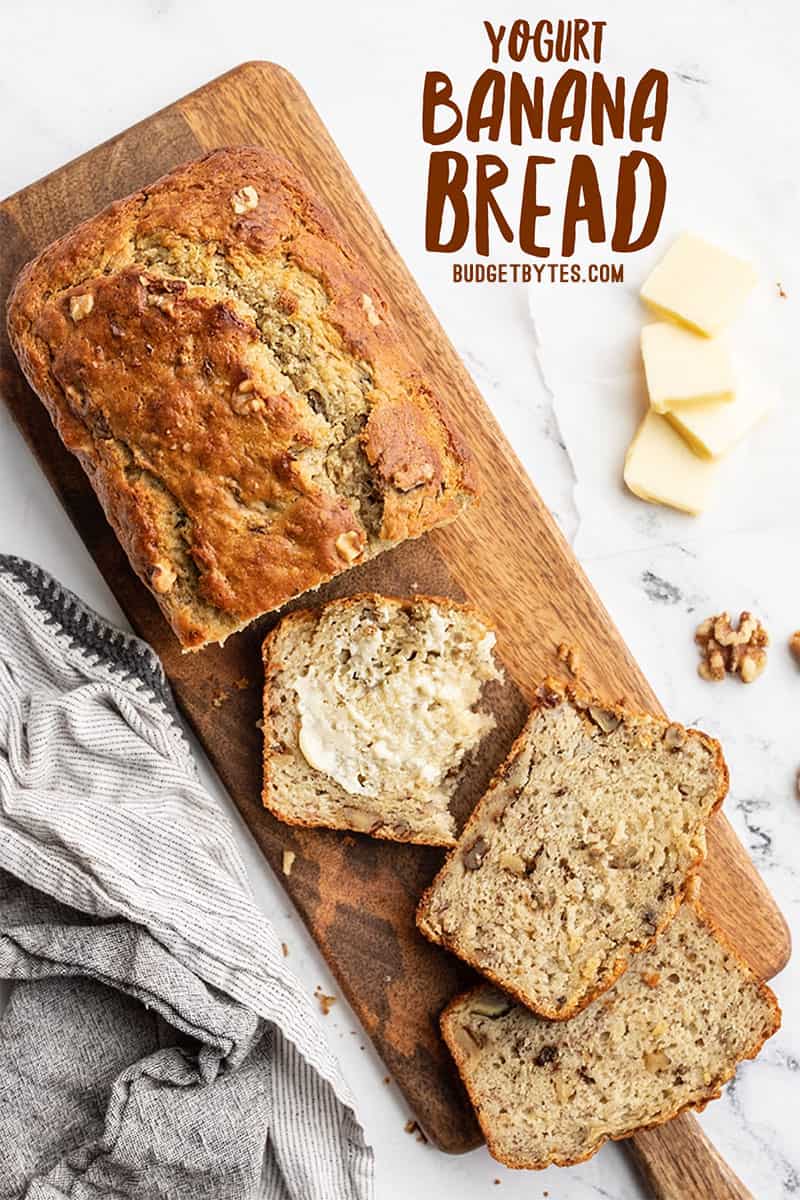 A half sliced loaf of yogurt banana bread on a wooden cutting board, with butter.