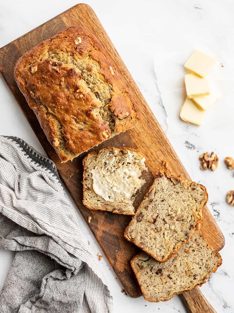 A loaf of banana bread on a narrow wooden cutting board, a few slices cut from the loaf and butter spread on one slice.