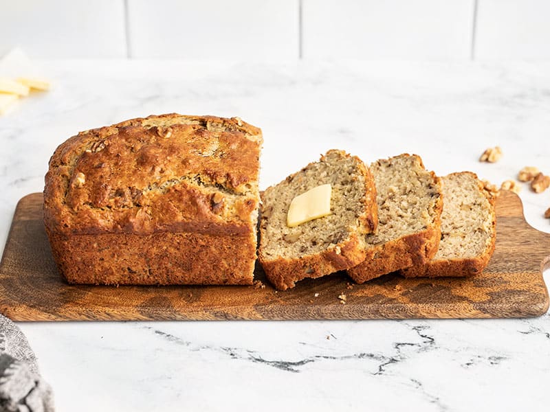 Side view of a loaf of yogurt banana bread on a wooden cutting board, half of it sliced, one slice with butter