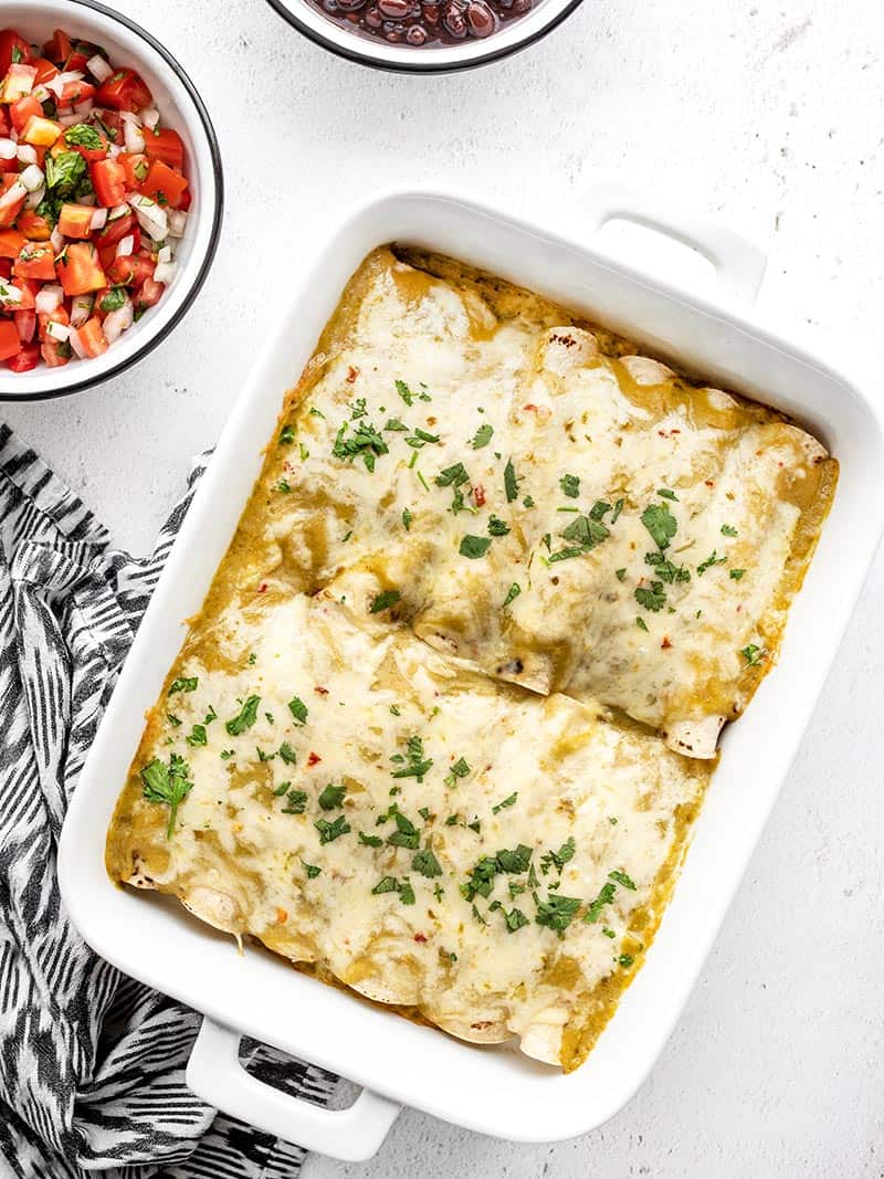 A casserole dish with green chile chicken enchiladas next to a bowl of pico de gallo