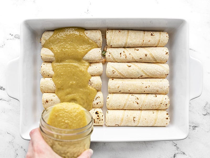 Green Chile Enchilada Sauce being poured from a jar onto a pan of rolled enchiladas from above