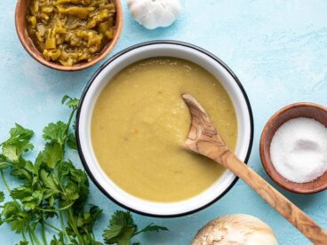 A wooden spoon in a bowl of green chile enchilada sauce with cilantro and diced green chiles on the side