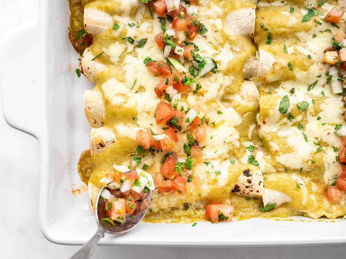 Pico de gallo being spooned over a casserole dish of green chile enchiladas.