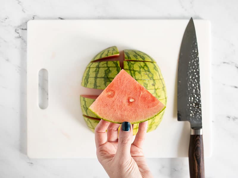 A hand holding a watermelon wedge with the rest of the watermelon in the background