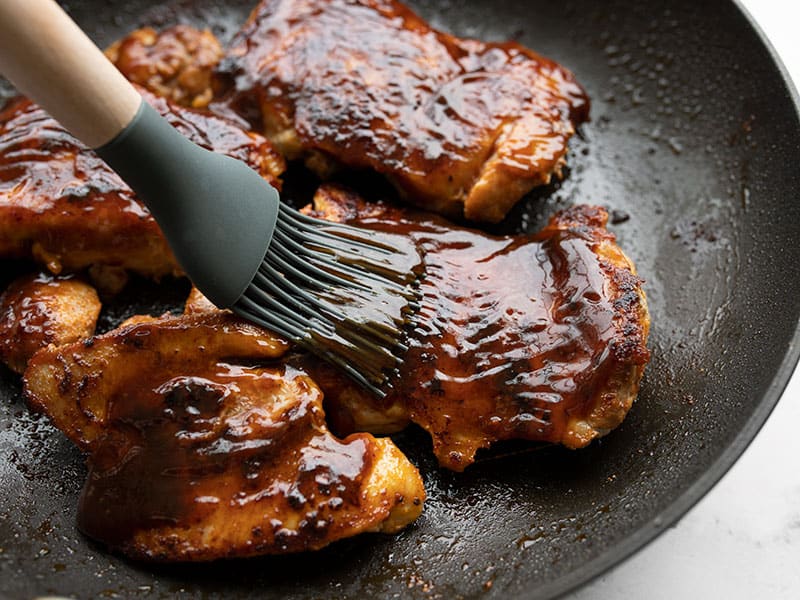 BBQ sauce being brushed onto chicken thighs in the skillet