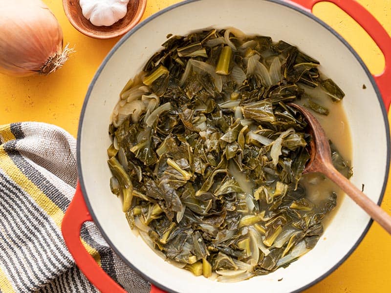 Jerk seasoned collard greens in a red pot, onion and garlic on the side