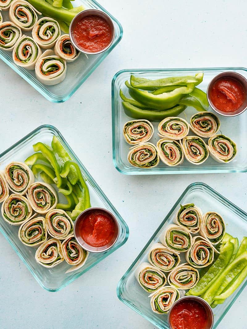 Four rectangular glass containers with pizza roll ups, green bell pepper, and a dipping cup of pizza sauce.