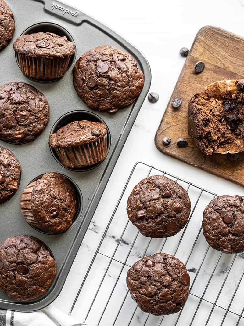 Chocolate banana muffins in the muffin tin, on a cooling rack, and a wooden cutting board