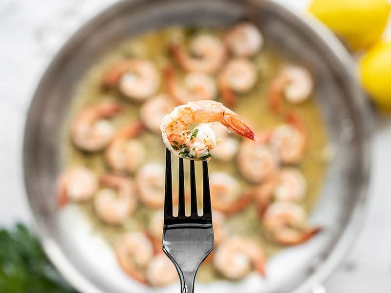 Close up of one garlic butter shrimp with the skillet full in the background