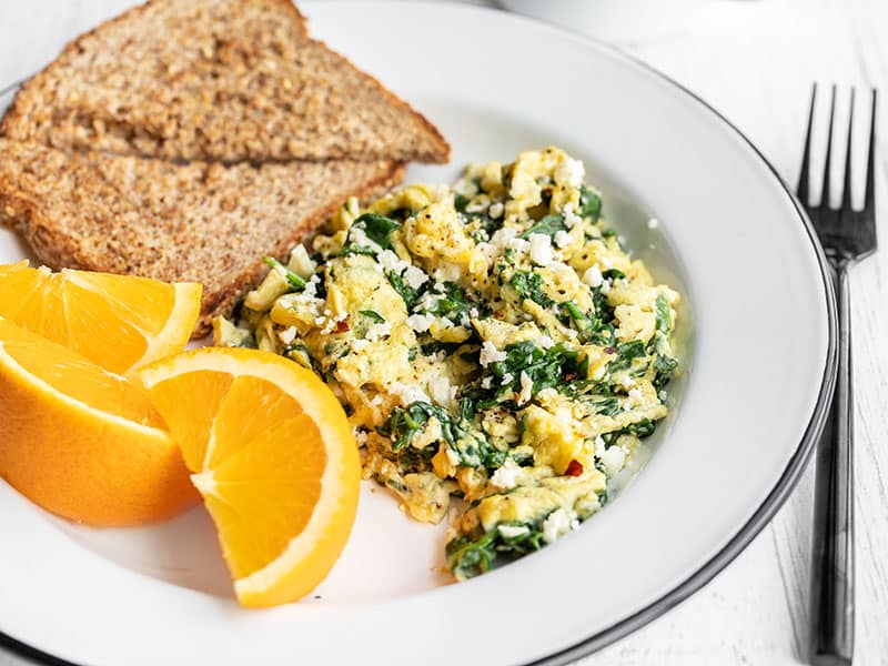 Side view of a plate with scrambled eggs with spinach and feta, toast, and oranges