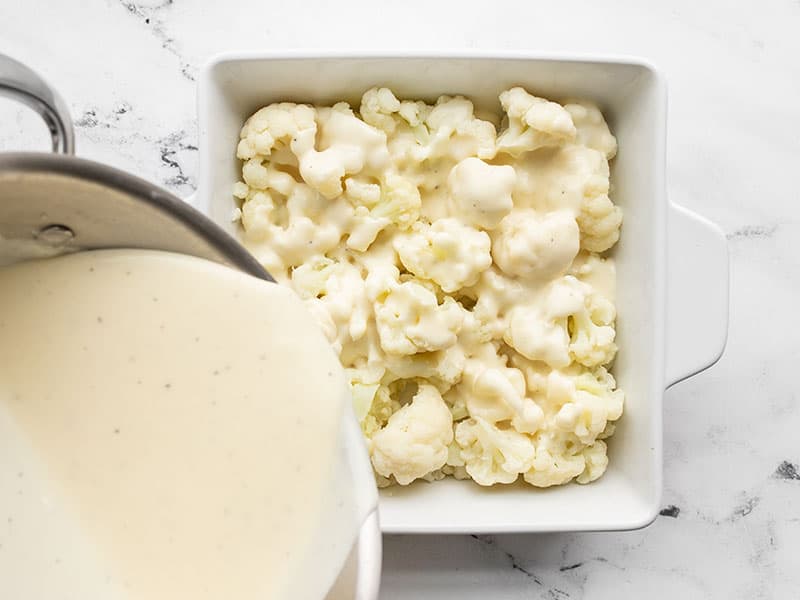 cheese sauce being poured over cauliflower in casserole dish