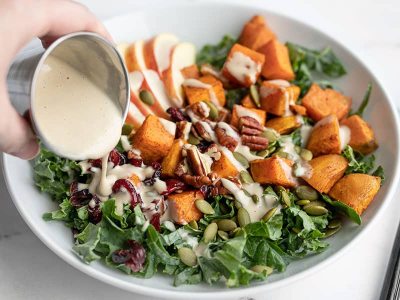 front view of maple tahini dressing being poured over autumn kale and sweet potato salad