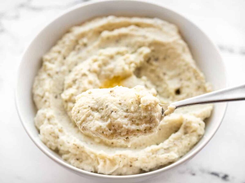 A spoonful of mashed cauliflower held close to the camera, the bowl in the back.