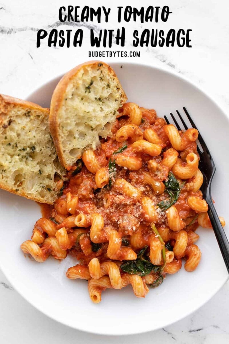 Overhead view of a bowl of creamy tomato pasta with sausage and garlic bread, title text at the top