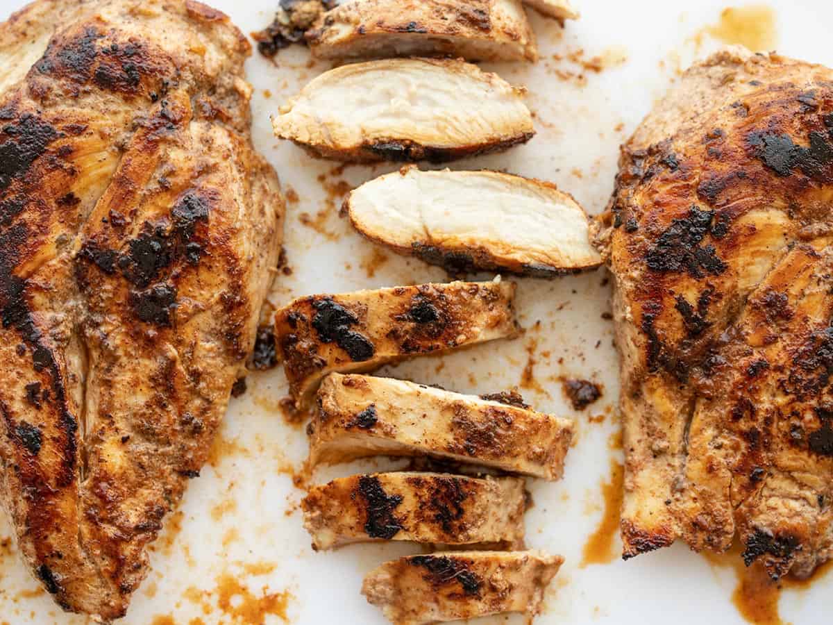 Sliced yogurt marinated chicken on a cutting board next to two whole cooked chicken breasts