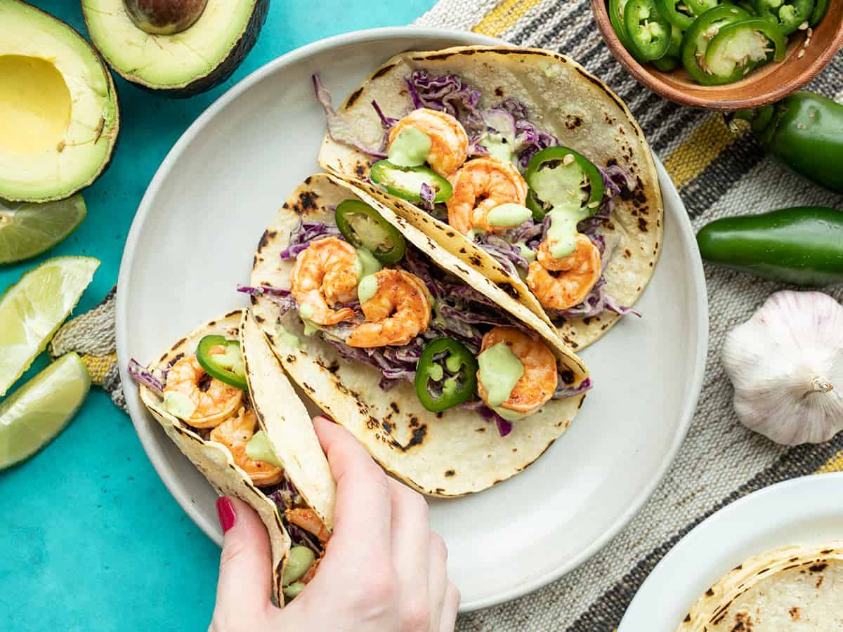 A hand picking up one Tajín Shrimp Taco off a plate of three