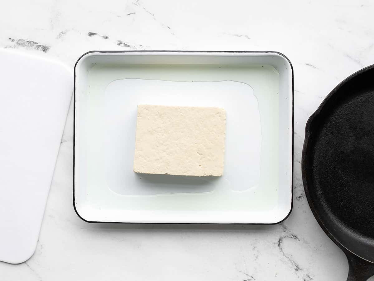 A block of tofu on a baking tray with a cast iron skillet on one side, cutting board on the other side