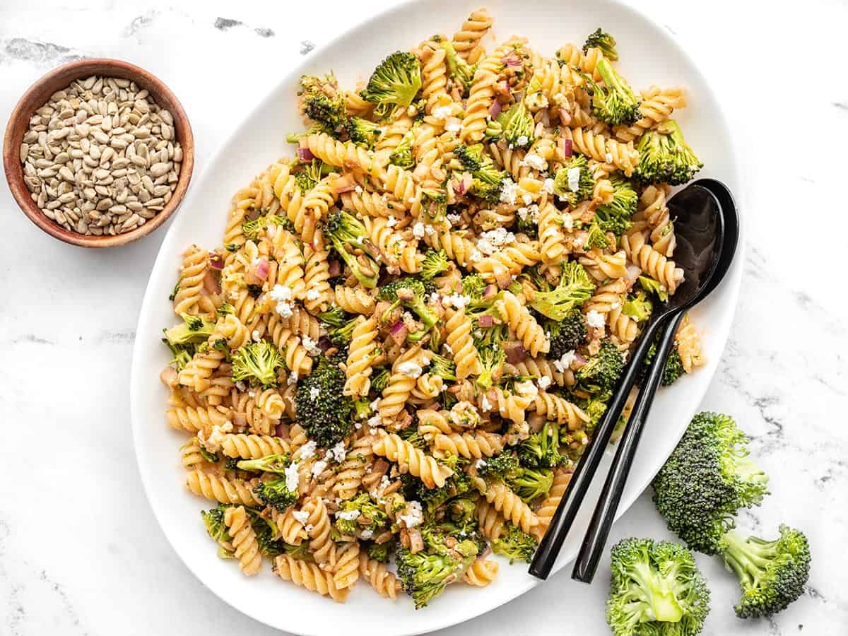 Overhead of broccoli pasta salad on a platter with a bowl of sunflower seeds and broccoli florets on the side