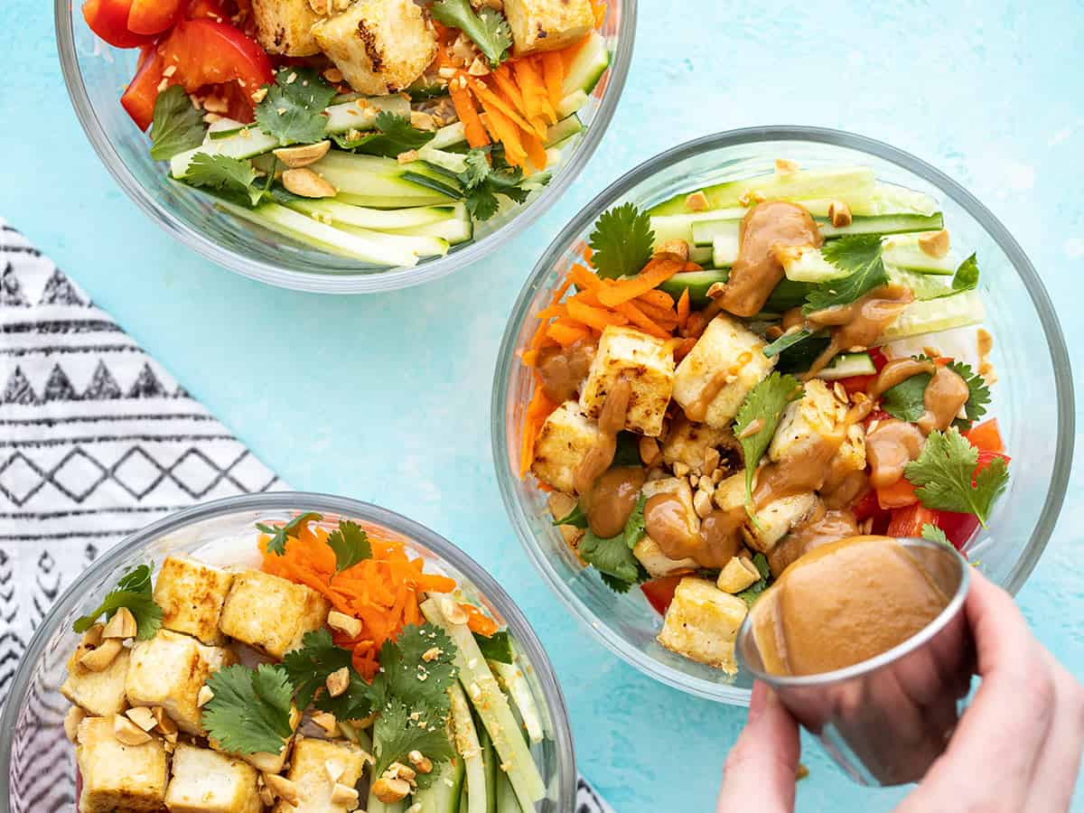 Three peanut tofu noodle bowls in glass containers, dressing being poured over one