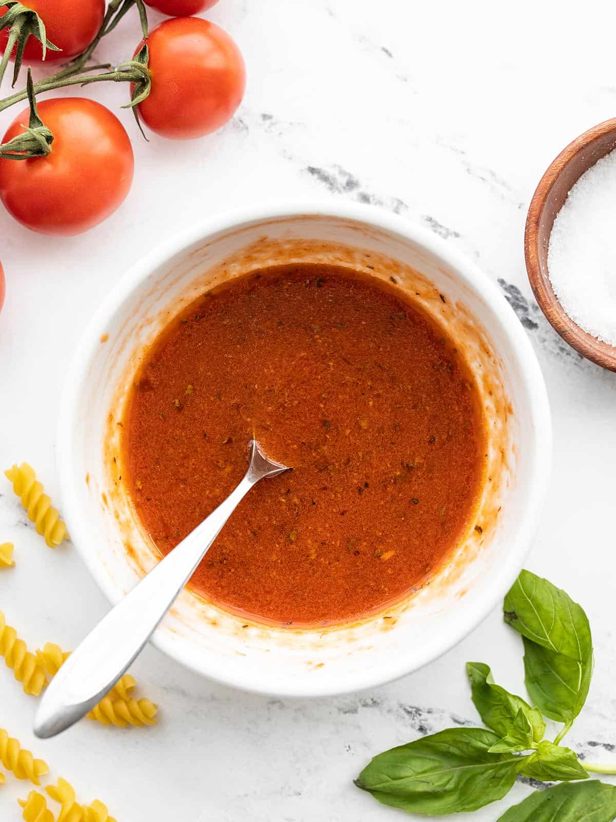 Overhead view of a bowl full of tomato basil vinaigrette with tomatoes, dry pasta, and spinach on the side