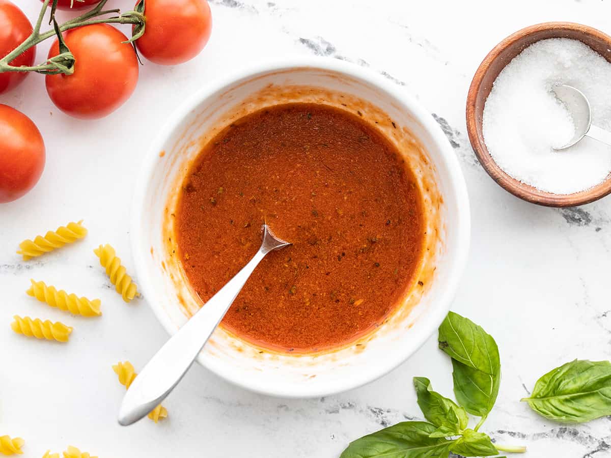 Overhead view of a bowl full of tomato basil vinaigrette with tomatoes, pasta, salt, and basil on the side of the bowl