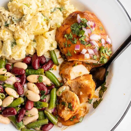 BBQ Cheddar Baked Chicken on a plate with potato salad and bean salad, a fork grabbing a slice of chicken