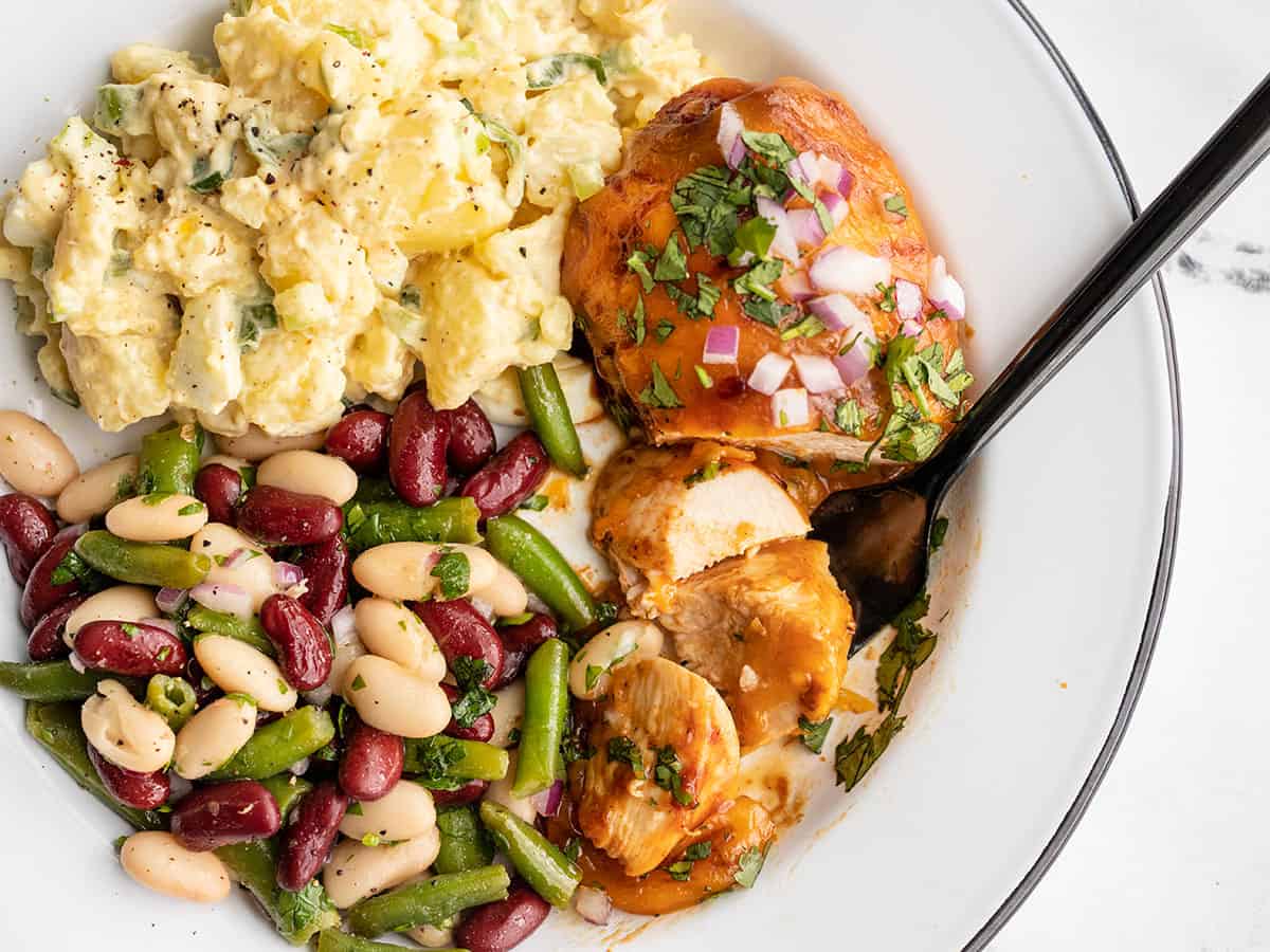 BBQ Cheddar Baked Chicken on a plate with potato salad and bean salad, a fork grabbing a slice of chicken