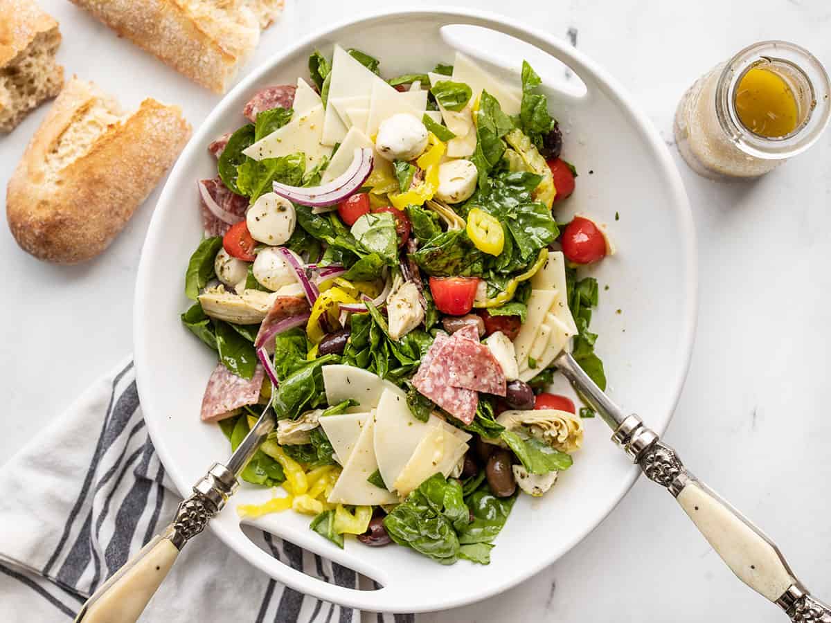 Salad tongs in a half stirred antipasto salad with bread and dressing on the side