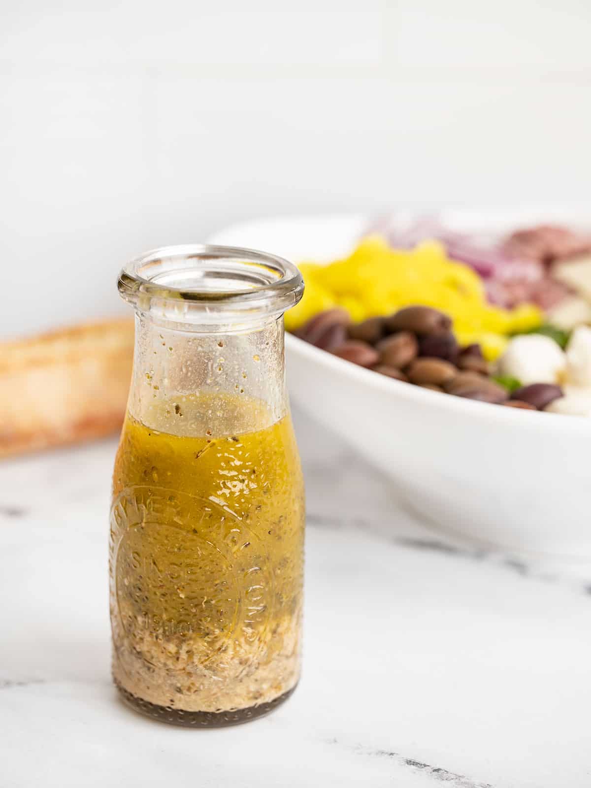 a small glass carafe with Italian dressing with a salad bowl in the back