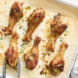 Overhead view of baked chicken drumsticks on a baking sheet, with one being removed with tongs.