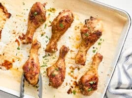 Overhead view of baked chicken drumsticks on a baking sheet, with one being removed with tongs.